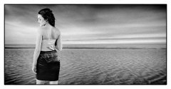 Foto gemaakt in greenscreen fotostudio in zwart-wit fotomodel poseert met zwart rokje en wit topje ze staat met haar rug naar de camera en kijkt opzij ze staat op het strand met de zee op de achtergrond gefotografeerd door fotograaf fotostudio Marijnissen Fotografie Rotterdam.