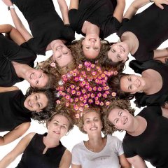 Vrijgezellenfeest fotoshoot met 9 dames in het zwart en de aanstaande bruid in het wit ze liggen op de grond met alle hoofden bij elkaar en kijken omhoog naar de camera tussenin zijn allemaal bloemetjes  gefotografeerd door fotograaf in de fotostudio Rotterdam  www.marijnissenfotografie.nl